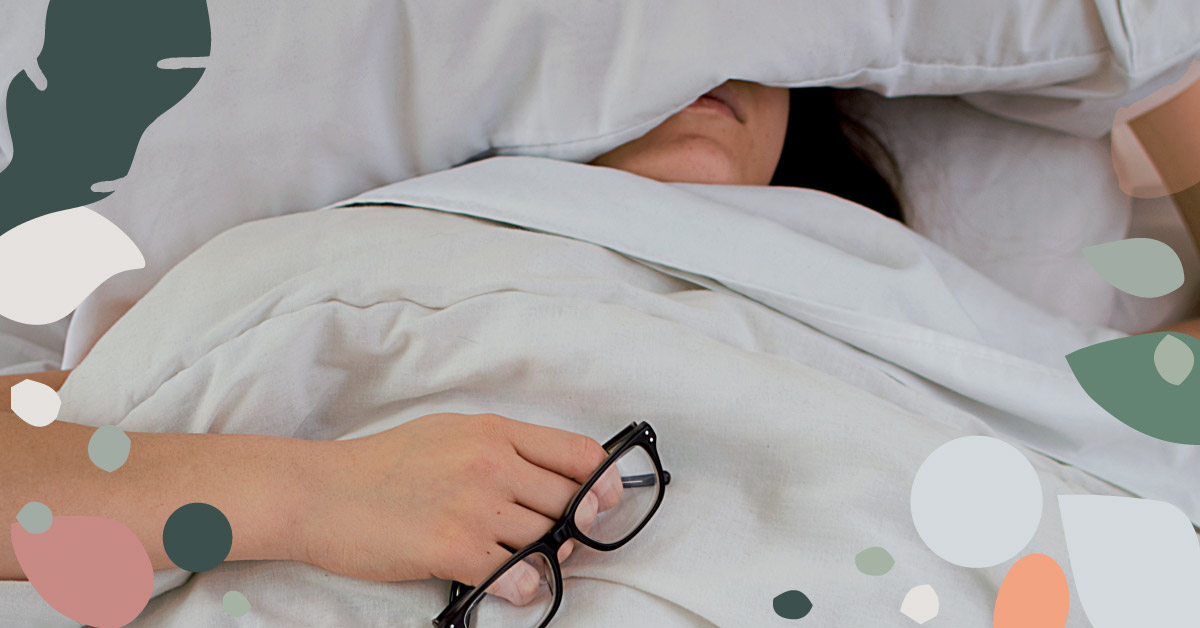 image of woman sleeping in bed, covering the top half of her head with a pillow while holding her glasses in the other hand